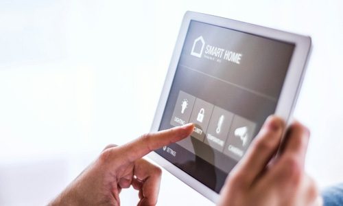 Hands holding a tablet device displaying smart home controls such as lighting, security, temperature and cameras. 